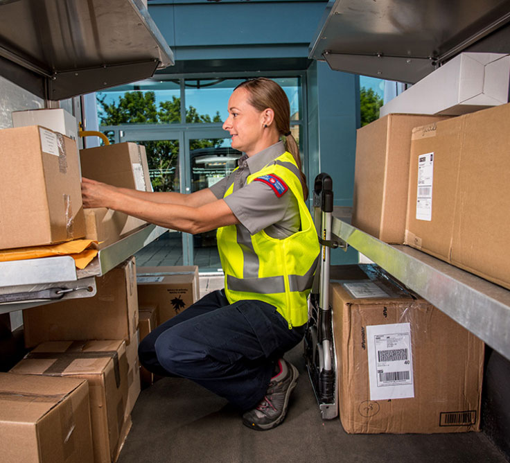 A Canada Post employee wears a yellow safety vest and crouches to reach for a parcel in the back of a Canada Post delivery vehicle.
