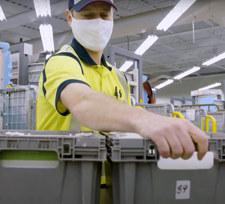 A Canada Post employee wears a yellow safety vest and a face mask. He grabs a bin of mail in a sorting facility.