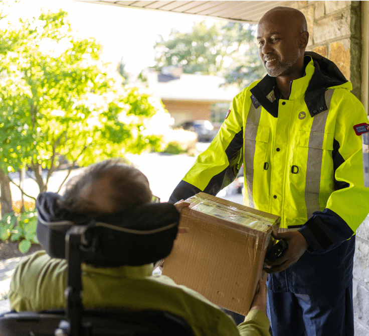 A Canada Post mail carrier hands a parcel to a man using a wheelchair on his front porch.