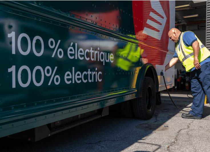 A Canada Post employee wearing a high-visibility yellow safety vest charges a Canada Post electric delivery truck.