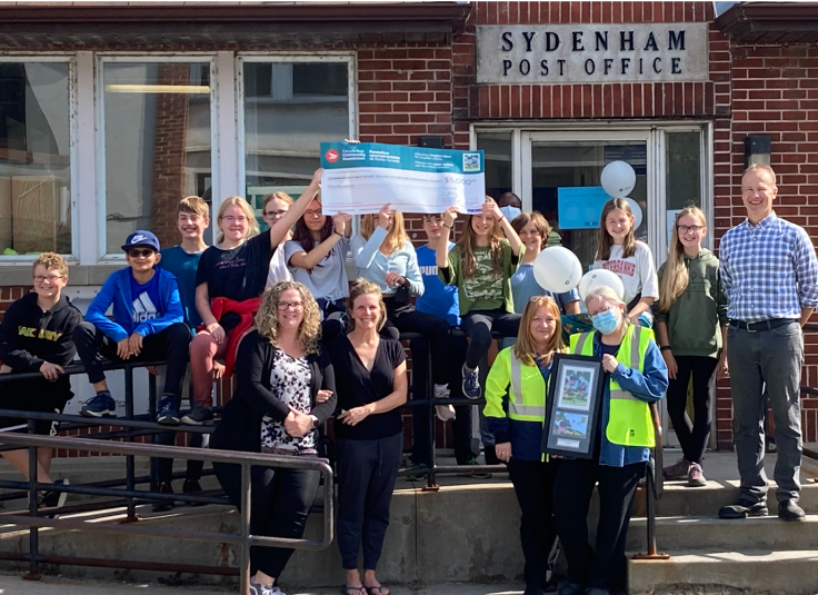 Des élèves et des membres du personnel de l’école publique Loughborough posent à l’extérieur du bureau de poste de Sydenham tenant un grand chèque de la Fondation communautaire de Postes Canada