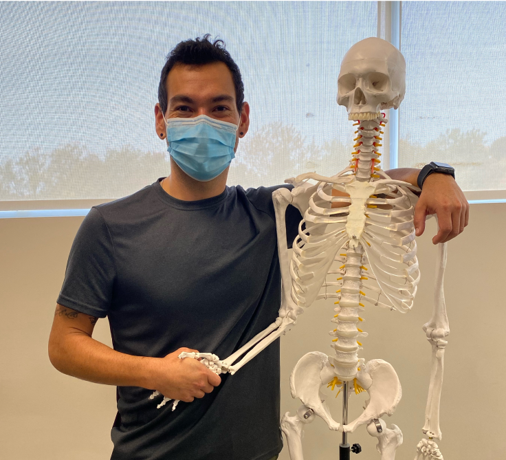 Chett Monague, a recipient of a Canada Post Award for Indigenous Students, wears a face mask and shakes hands with a display skeleton.