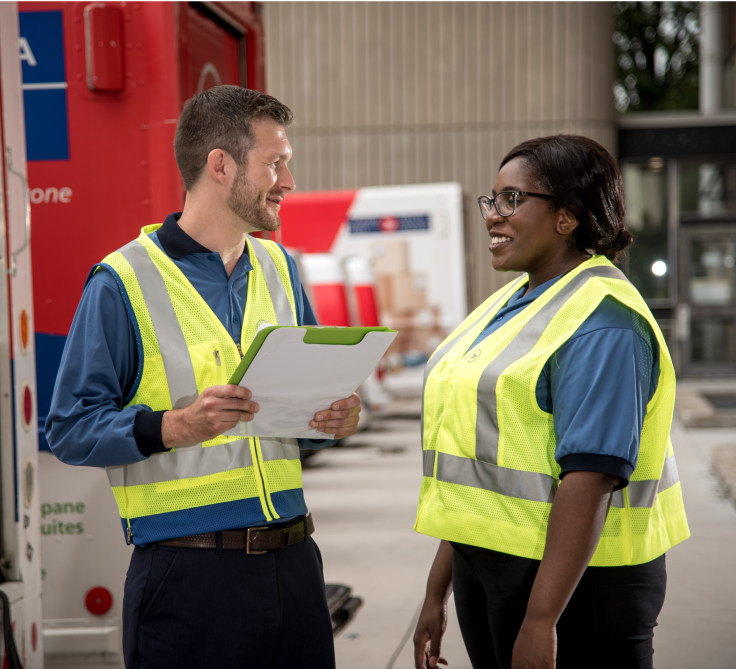 Two Canada Post employees wearing bright yellow vests talk to each other in a Canada Post facility. 