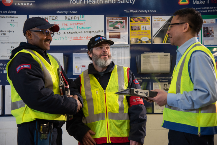 A man in a blue shirt and a yellow safety vest holds a clipboard and talks to two men in black jackets and yellow safety vests in a Canada Post facility.