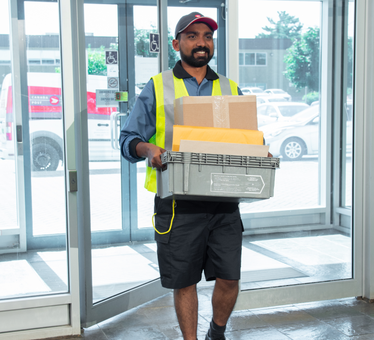 Un employé de Postes Canada vêtu d’un gilet de haute visibilité jaune tient une boîte remplie de courrier en entrant dans un immeuble par des portes vitrées