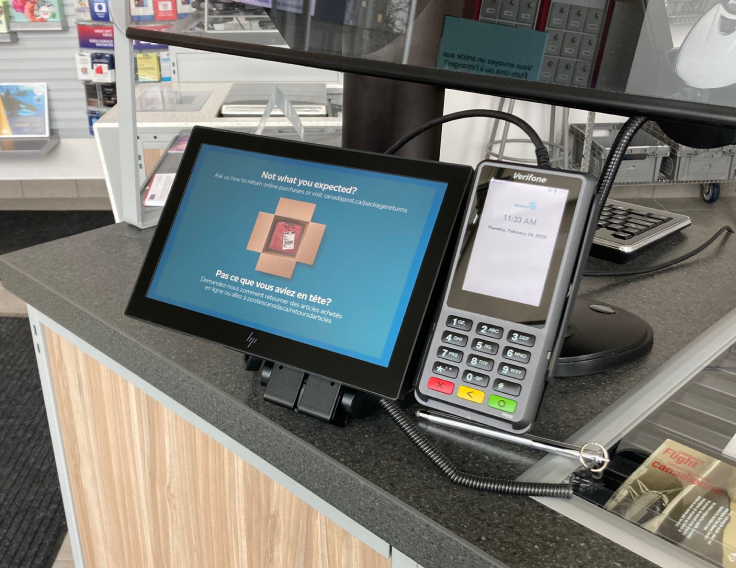 A customer-facing touchscreen and a mobile hand-held payment and signature device at a Canada Post post office. The touchscreen device displays an open shipping box.