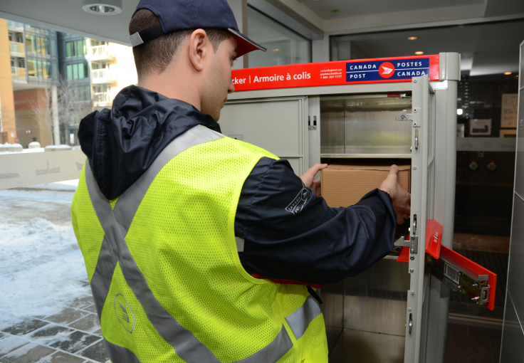 A Canada Post letter carrier wearing a high visibility yellow vest deposits a parcel into a parcel locker at a community mailbox in winter.