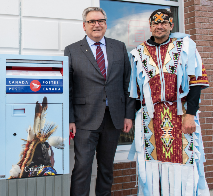 Canada Post President and CEO Doug Ettinger and David Meuse, cultural ambassador for the Membertou Trade and Convention Centre, pose together beside a mailbox outside the Canada Post Membertou post office. Meuse wears colourful, traditional Mi'kmaq attire.