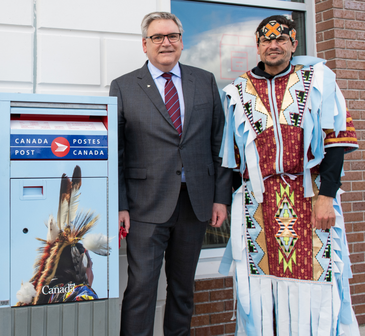 Doug Ettinger, président-directeur général de Postes Canada, et David Meuse, ambassadeur culturel du Membertou Trade and Convention Centre, posent à côté d’une boîte aux lettres au bureau de poste de Membertou. David Meuse porte une tenue micmaque traditionnelle colorée