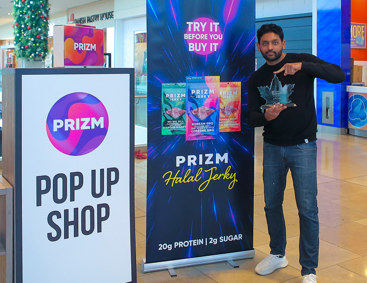 Zeeshan Hayat, co-founder of PRIZM Foods, holds his Tales of Triumph Contest award and stands beside a pop up banner advertising his brand in a pop up shop location.