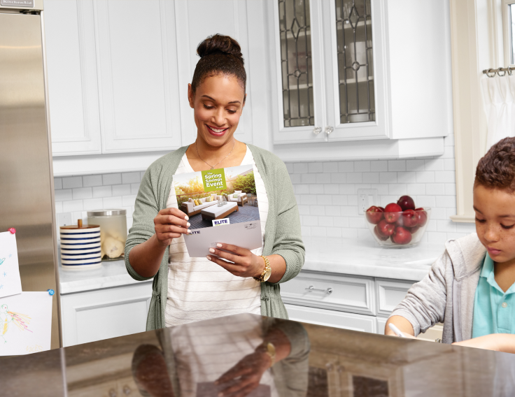A woman smiles as she reads a flyer in her kitchen. Her child stands nearby.