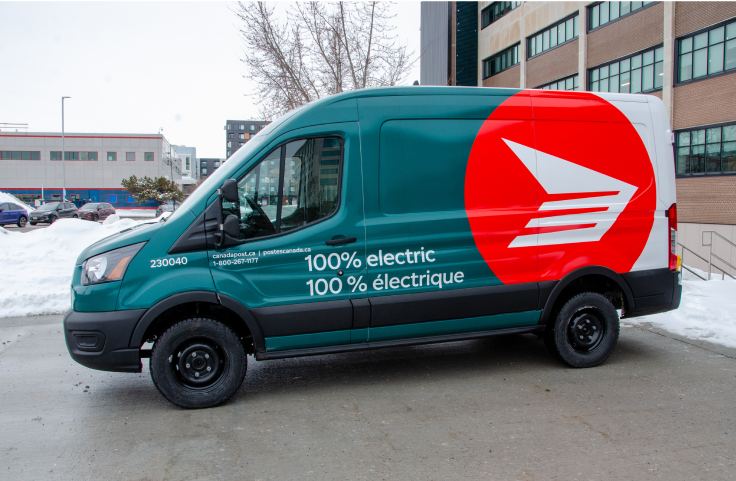 A Canada Post green fleet electric delivery vehicle is parked outside a Canada Post facility. It says