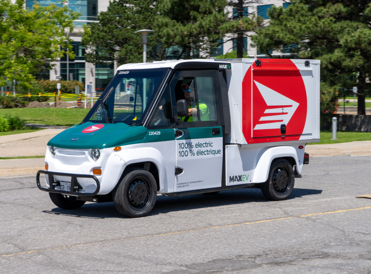 Un nouveau véhicule électrique à basse vitesse de Postes Canada roule sur une rue. Il est vert forêt et blanc, et porte l’icône du logo rouge de Postes Canada