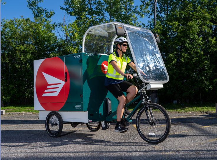 Une cycliste portant des vêtements de haute visibilité jaunes se déplace sur un vélo cargo électrique de Postes Canada vert forêt et blanc, orné de l’icône du logo rouge de l’entreprise