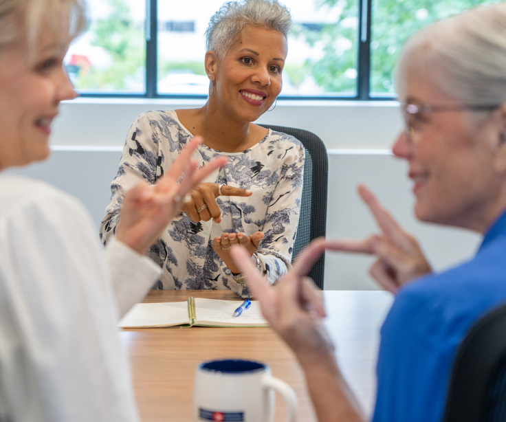 A grey-haired woman signs to a blonde woman and a grey-haired woman in a boardroom.