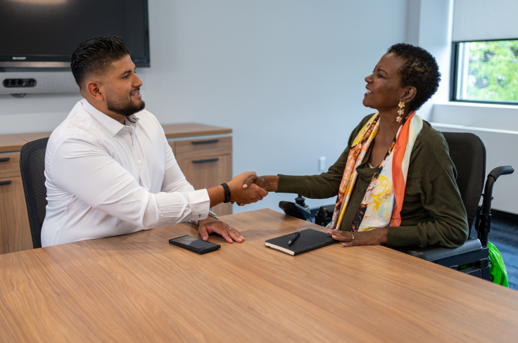 A woman with a notebook using a wheelchair at a boardroom table and shaking hands with a man.