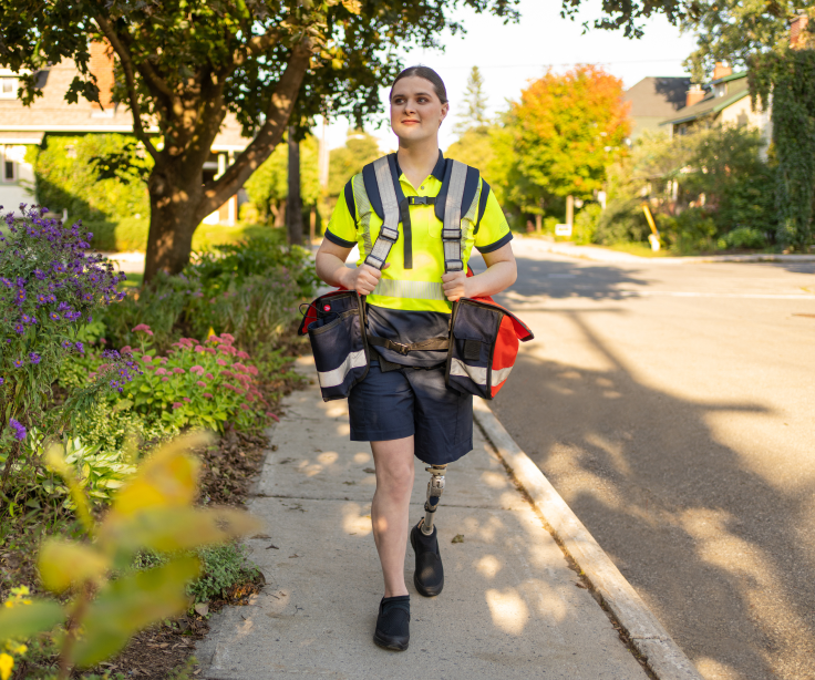 A Canada Post mail carrier wearing saddlebags smiles and walks down a residential street.