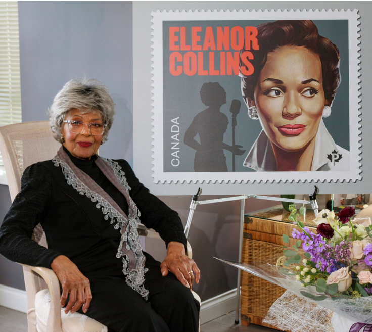 Eleanor Collins sits and poses for a photo at a virtual event in which her commemorative stamp was unveiled. She sits beside an enlargement of the stamp and a pastel flower arrangement.