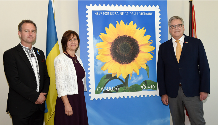 Orest Sklierenko et Oksana Kuzyshyn de la Fondation Canada-Ukraine posent avec Doug Ettinger, président-directeur général de Postes Canada, à Ottawa, lors du dévoilement du timbre philanthropique Aide à l’Ukraine. Un agrandissement du timbre les sépare