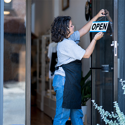 A woman wearing a black apron stands in the doorway of a storefront, hanging an "OPEN" sign on a glass door.