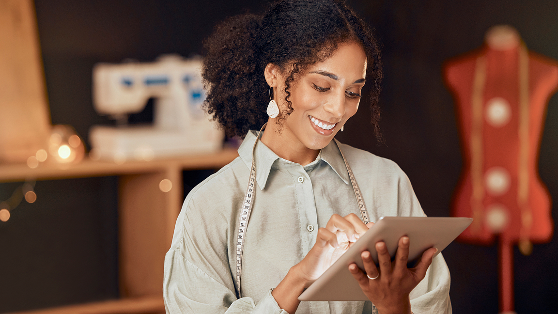 Smiling small business owner in light shirt with measuring tape around neck uses tablet in sewing workspace. Smiling small business owner in light shirt with measuring tape around neck uses tablet in sewing workspace.