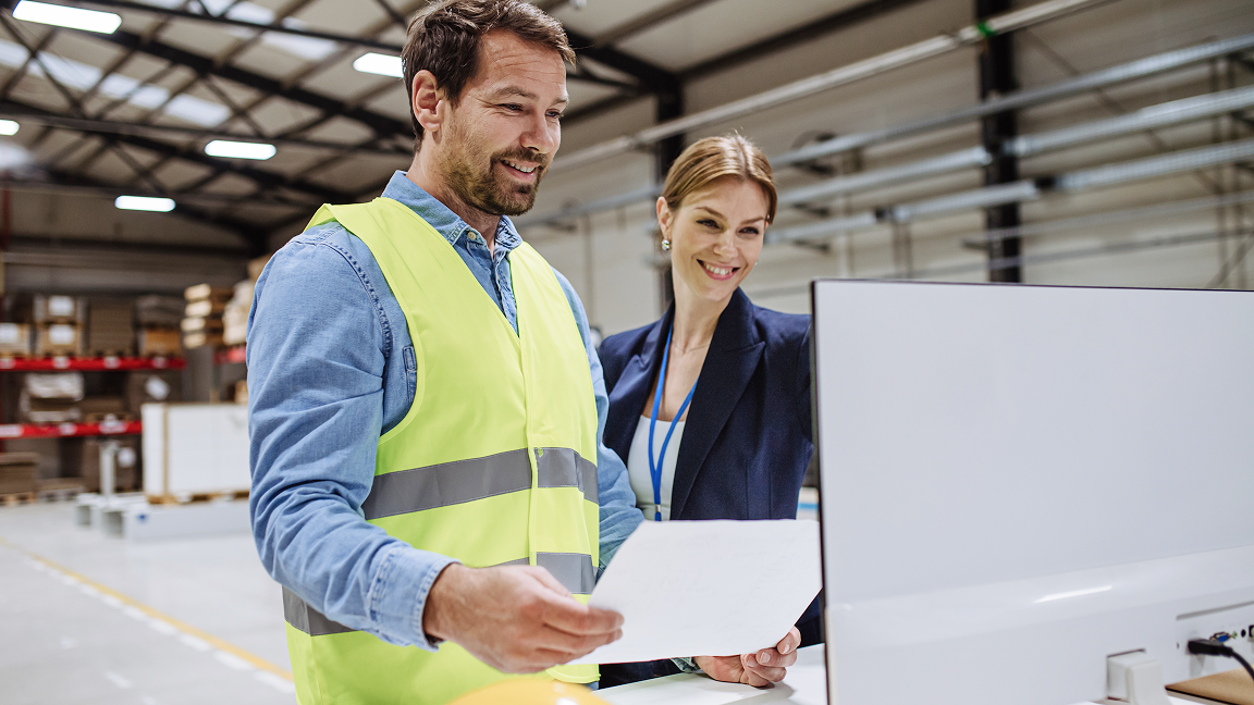 Two people reviewing documents together in an industrial workspace, with one wearing a high‑visibility vest. Two people reviewing documents together in an industrial workspace, with one wearing a high‑visibility vest.