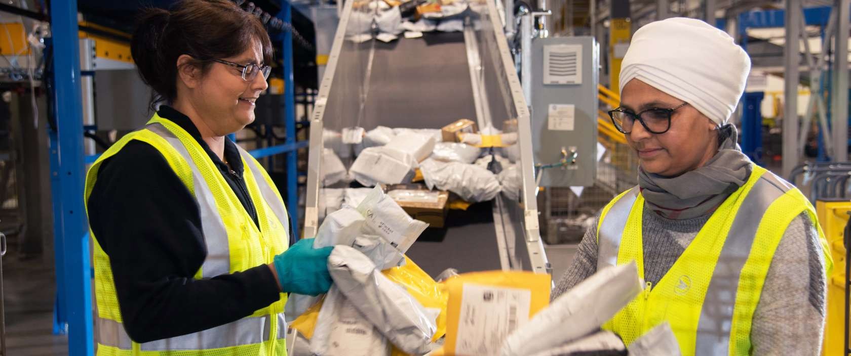 Two female Canada Post employees wearing bright yellow safety vests sort through packages and envelopes.