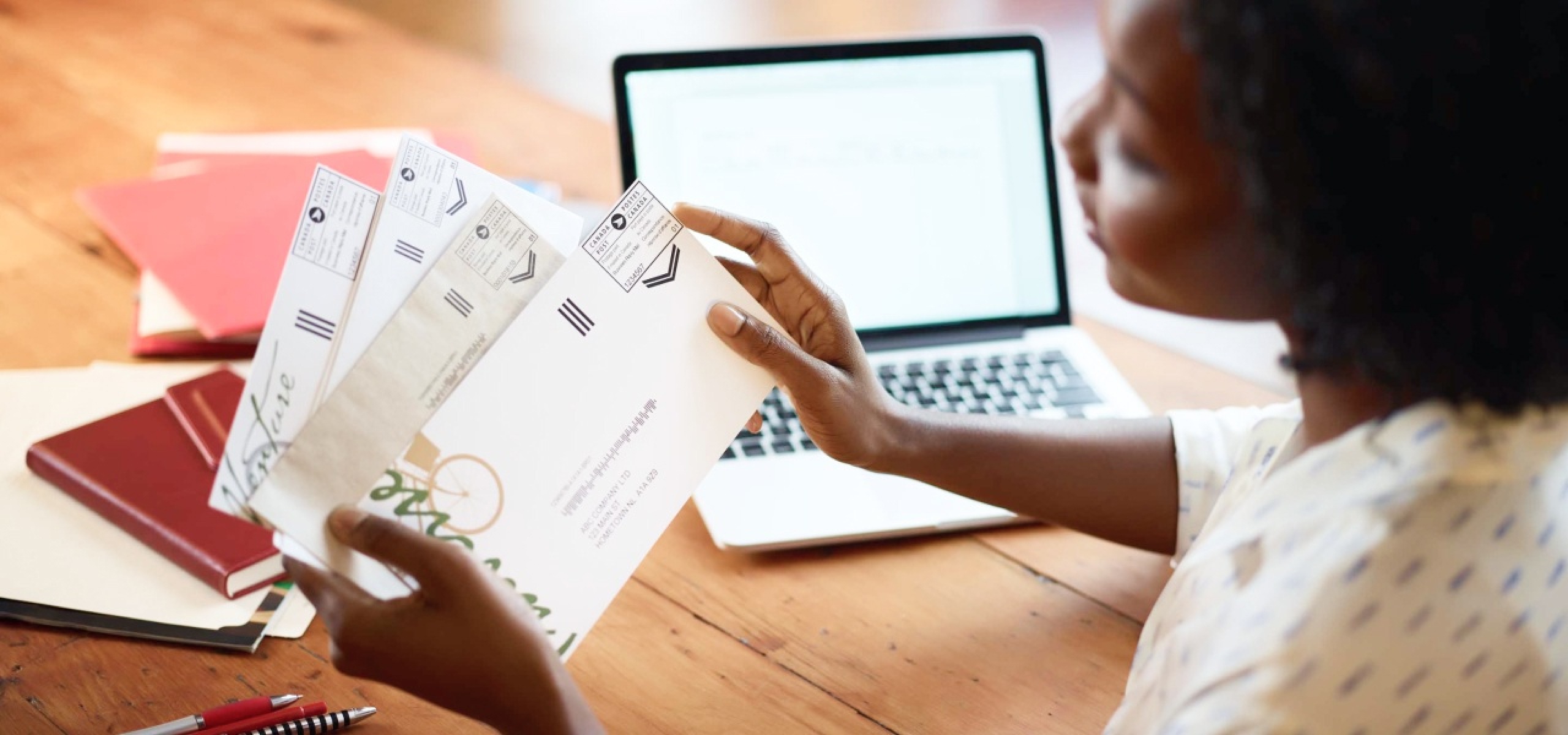 A woman at her desk holds a small stack of business mail. A Canada Post postal indicia is printed on the upper right-hand corner of each envelope.