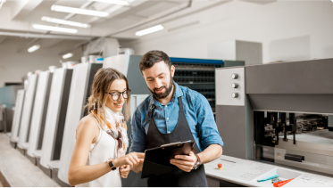 Two people consult a clipboard with large printing machinery in the background.