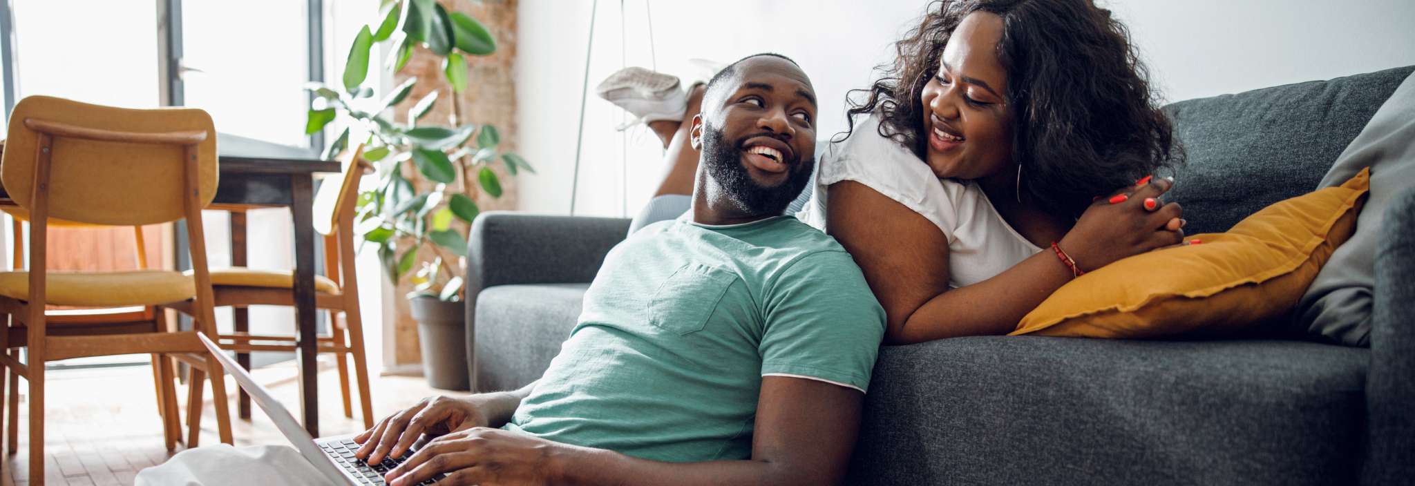A man types on a laptop and leans against a couch a woman is sitting on. They smile at each other.