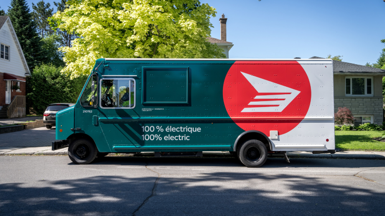 A new Canada Post truck drives on a residential street. The truck is forest green and white and features the red and white Canada Post rondelle logo.