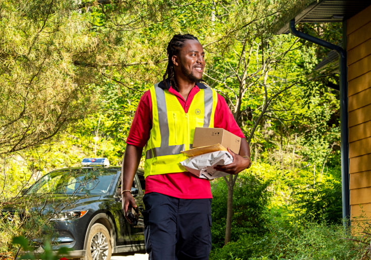 A mail carrier in a safety vest holds multiple parcels and walks through a treed residential area.