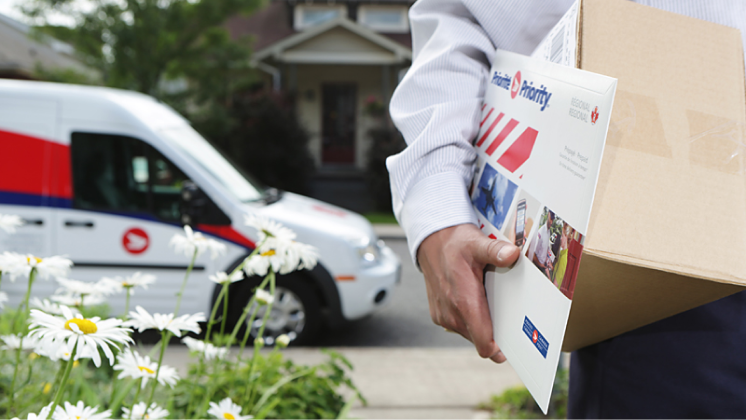 A letter carrier holds a shipping box and a bubble mailer. Their Canada Post vehicle is parked nearby.