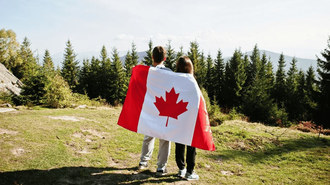 A man and woman wrapped in a Canadian flag look out over a forested area from a hilltop. A man and woman wrapped in a Canadian flag look out over a forested area from a hilltop.