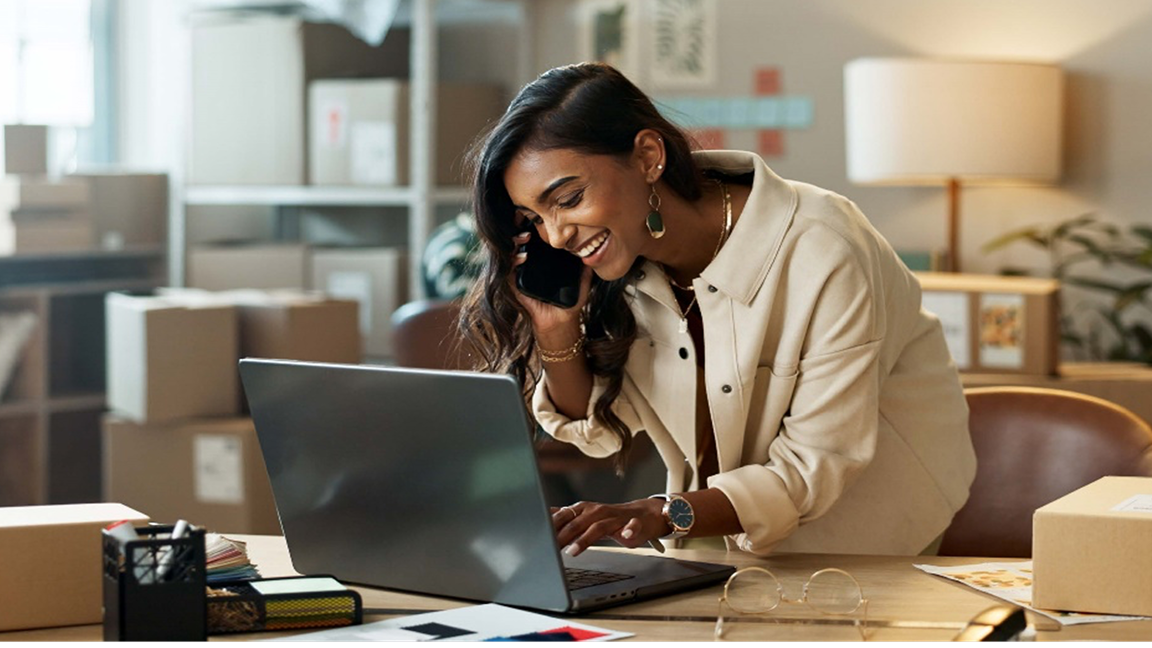A woman speaking on her cellphone smiles and types on a laptop in a room full of shipping boxes. A woman speaking on her cellphone smiles and types on a laptop in a room full of shipping boxes.