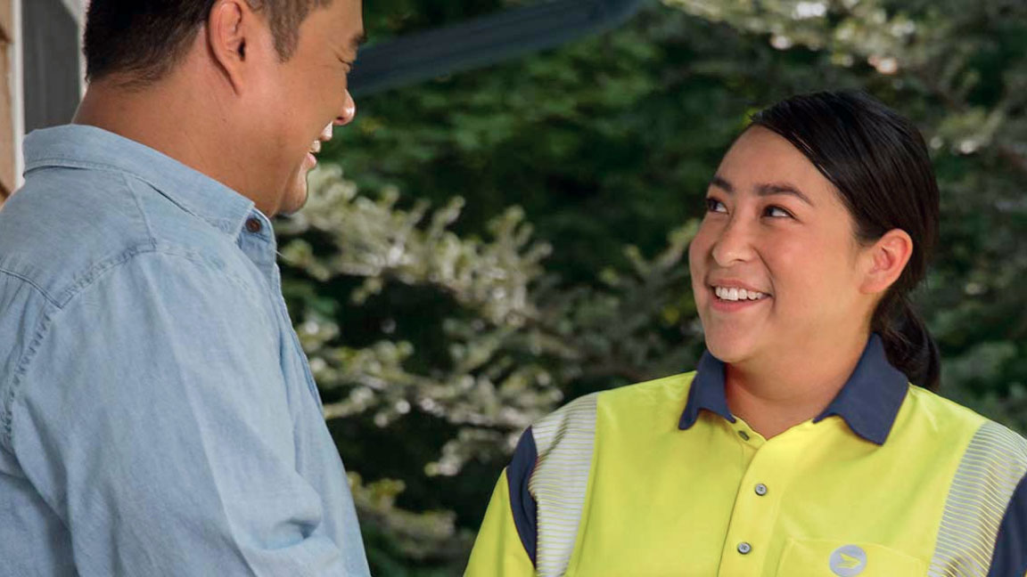 A smiling Canada Post mail carrier wearing a bright yellow jacket speaks with a customer on his front porch.