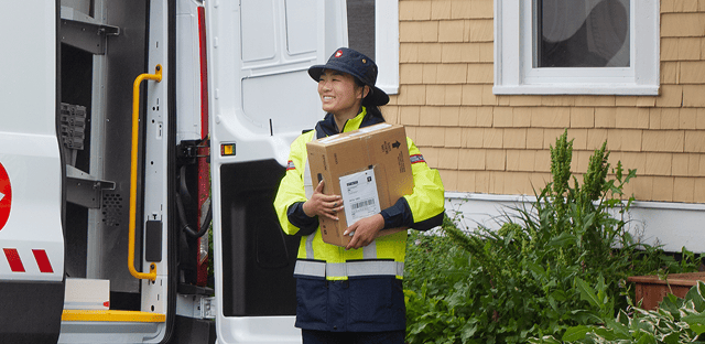 A postal worker in a high visibility uniform carries a large package while standing beside an open delivery vehicle outside a house.