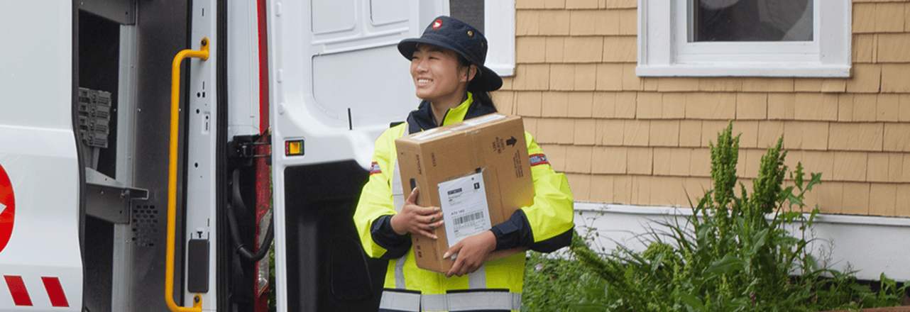 A postal worker in a high visibility uniform carries a large package while standing beside an open delivery vehicle outside a house.