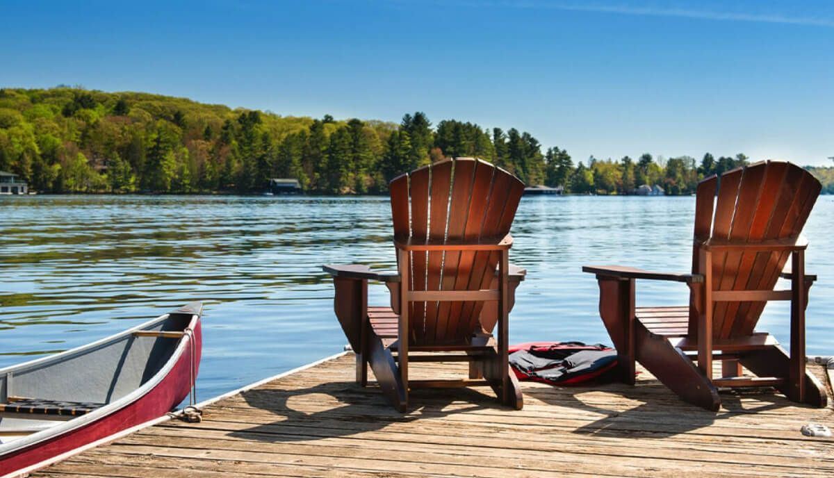 Two wooden chairs sit on a lakeside dock, next to a canoe.