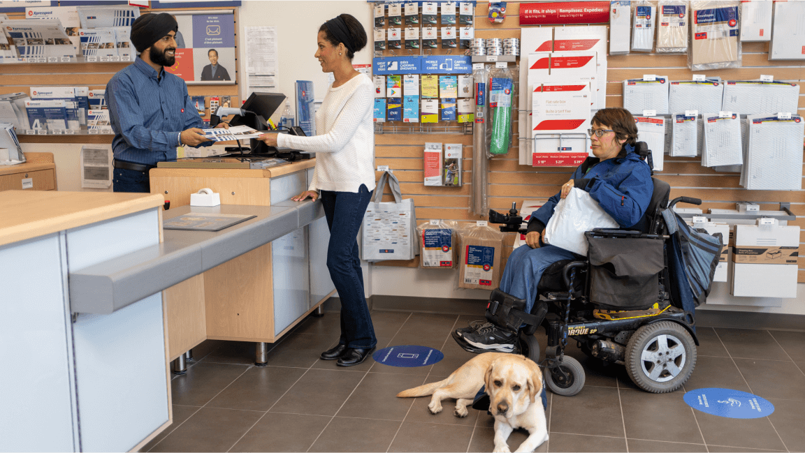A woman speaks to an employee at the desk of a post office. A wheelchair user with a service dog waits in line behind her in line.