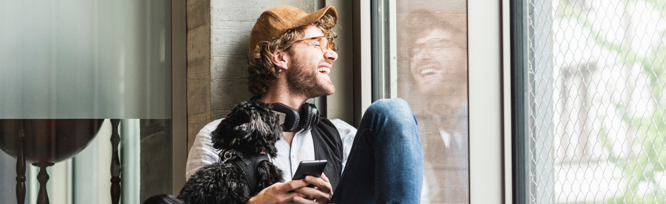 A young man laughs as he looks out a window. He holds a smart phone in his hands and cradles a dog in his arm.