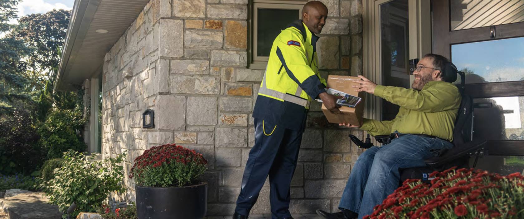 Un agent de livraison de Postes Canada tend un colis à un homme en fauteuil roulant devant sa porte d’entrée.