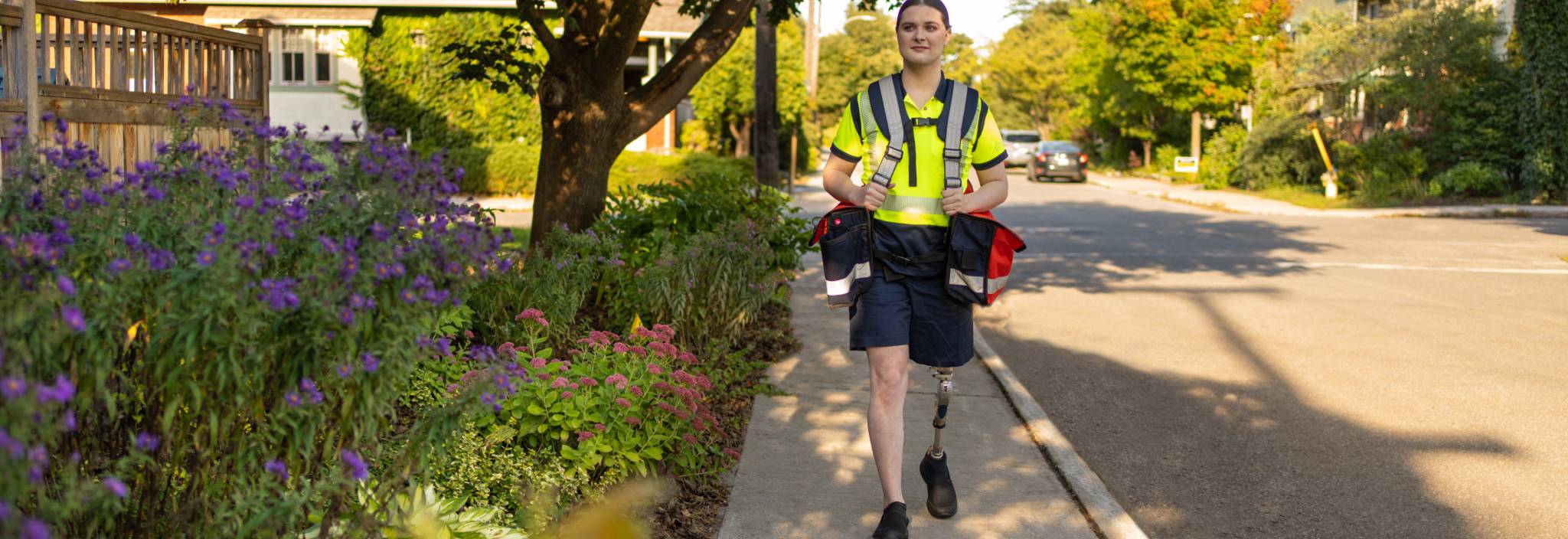 Une factrice de Postes Canada avec une jambe prothétique marche dans une rue résidentielle en portant des sacs de courrier. 