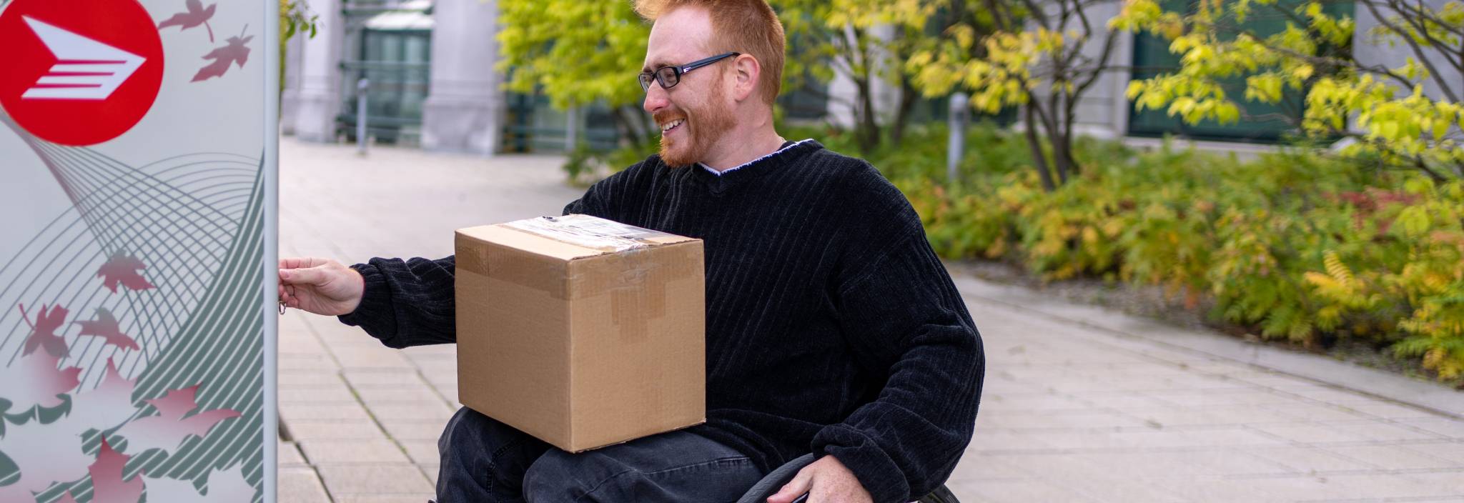 Un homme en fauteuil roulant ramasse un colis dans une boîte postale communautaire de Postes Canada.