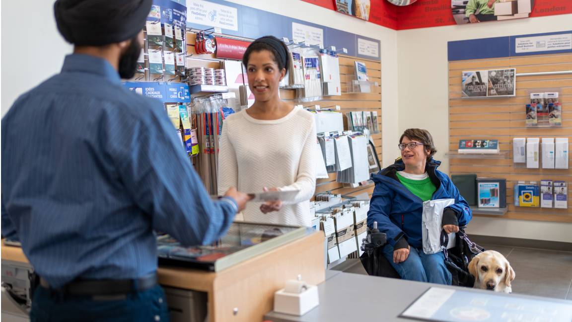 Un employé de bureau de poste interagit avec une cliente. Une femme en fauteuil roulant avec chien d’assistance fait la file. Un employé de bureau de poste interagit avec une cliente. Une femme en fauteuil roulant avec chien d’assistance fait la file.