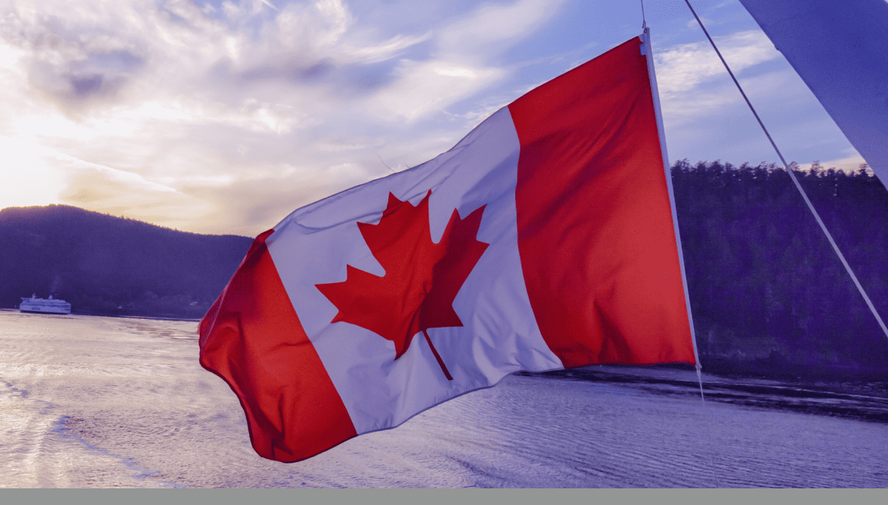 Un drapeau canadien flottant au vent, avec un plan d’eau et des collines boisées à l’arrière-plan.