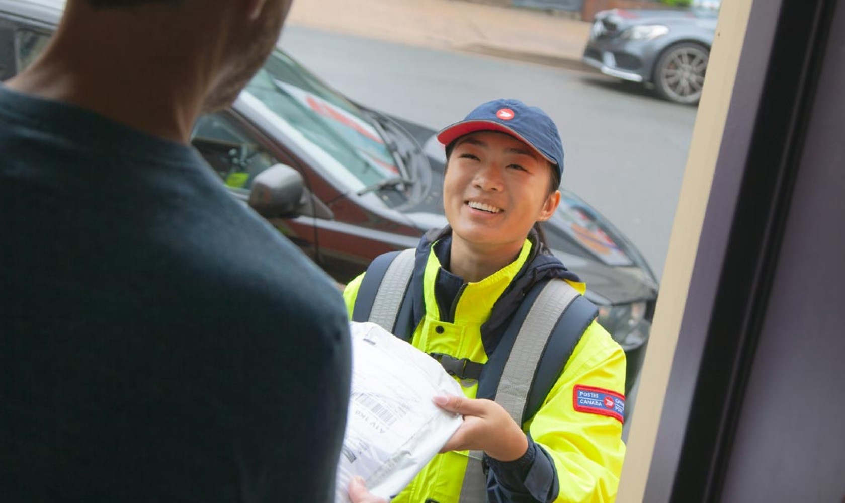 Une employée de Postes Canada portant un blouson jaune vif livre un colis à un homme debout dans l’entrée de sa maison.