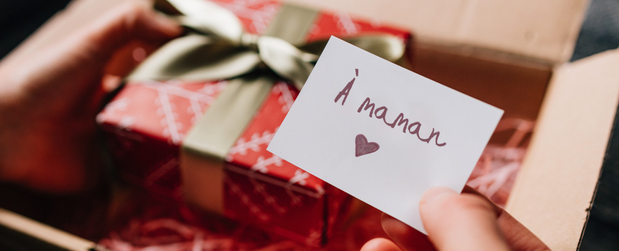 Des mains ouvrent un colis qui contient une boîte-cadeau emballée et une carte sur laquelle il est écrit « À maman »