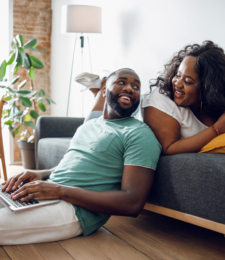 Un homme tape sur un ordinateur portable et s’appuie sur un canapé sur lequel une femme est assise. Les deux se sourient. Un homme tape sur un ordinateur portable et s’appuie sur un canapé sur lequel une femme est assise. Les deux se sourient.
