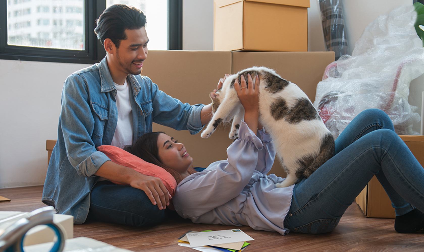 Un couple souriant joue avec un chat par terre. Il y a une pile de courrier à côté d’eux et des boîtes en carton empilées en arrière-plan.